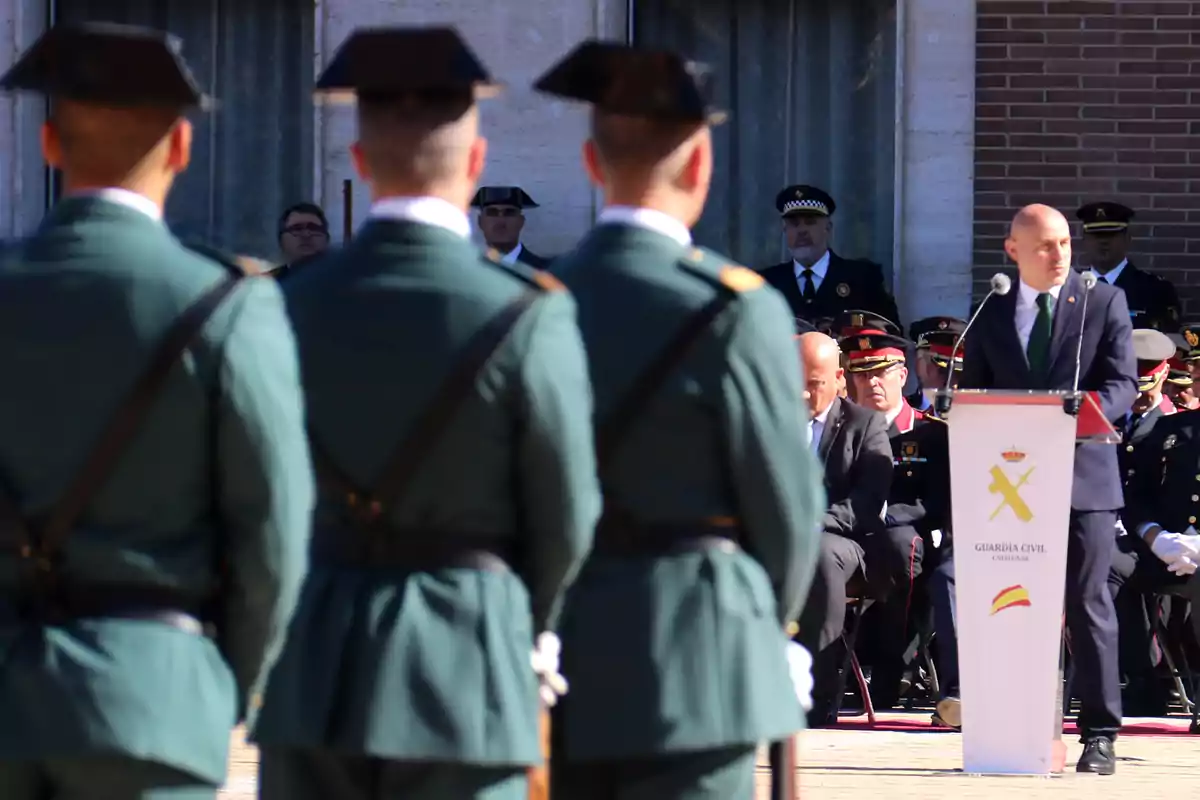 Un grup de guàrdies civils en uniforme verd assisteixen a un esdeveniment oficial a l'aire lliure on un home en vestit parla des d'un podi.