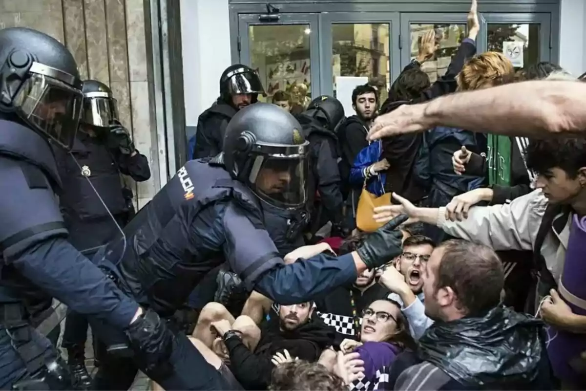 Policies antiavalots amb cascos i escuts enfrontant-se a un grup de manifestants en un carrer, alguns dels quals estan a terra mentre altres aixequen les mans.