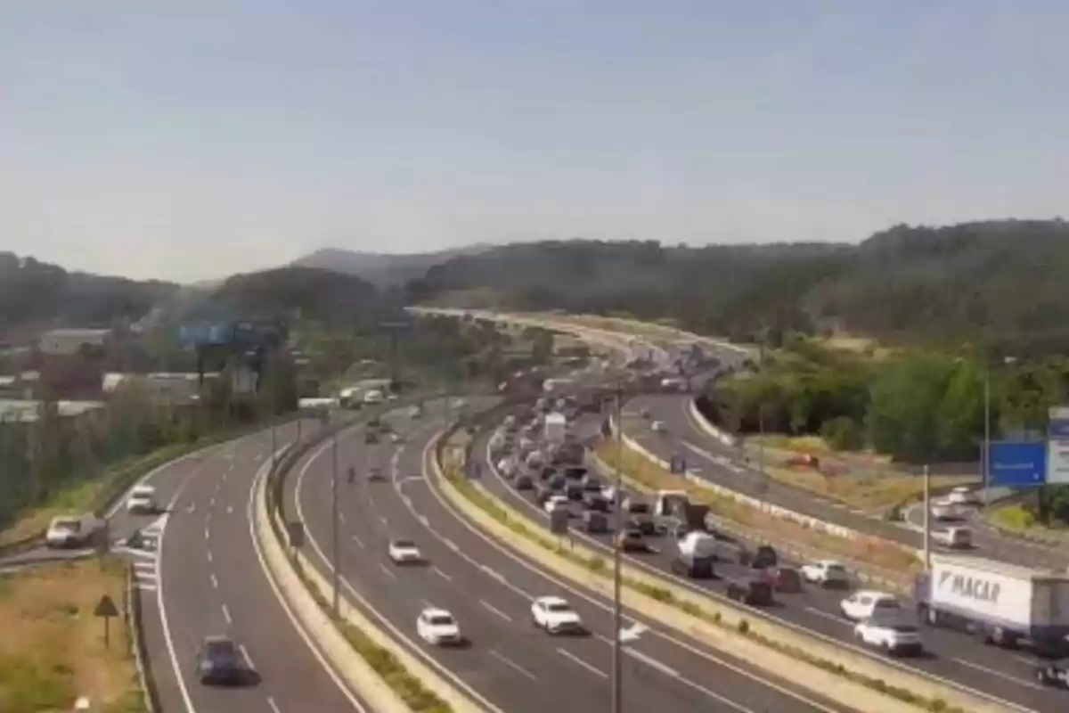 Autopista amb trànsit dens en un sentit i circulació fluida en l’altre, envoltada de vegetació i turons al fons