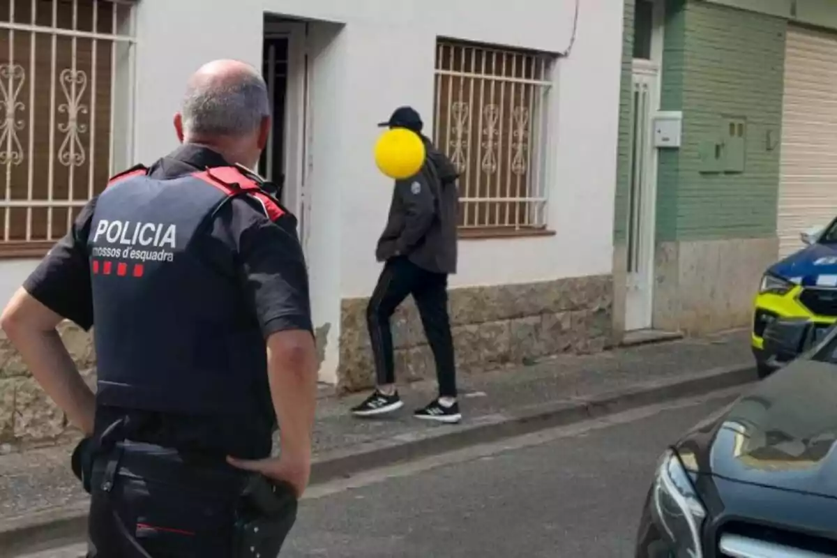 Un agent de policia observa una persona amb la cara pixelada que camina cap a l?entrada d?una casa en un carrer residencial.