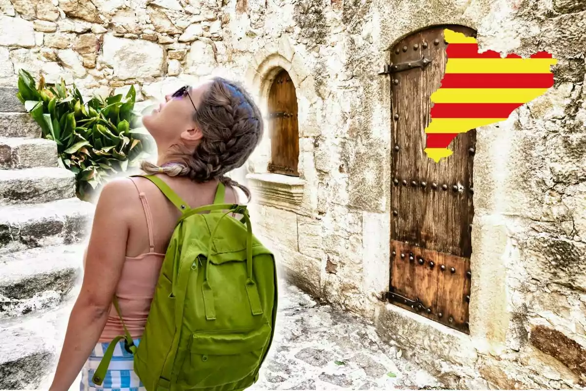 Una mujer con mochila verde y trenzas observa hacia arriba en una calle empedrada con una puerta de madera y una planta a su lado, con un mapa de Cataluña en la pared.