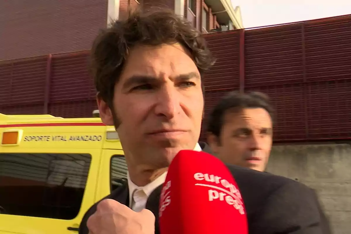 Cayetano Rivera con un micrófono de prensa roja frente a una ambulancia amarilla. Cayetano Rivera con un micrófono de prensa roja frente a una ambulancia amarilla.
