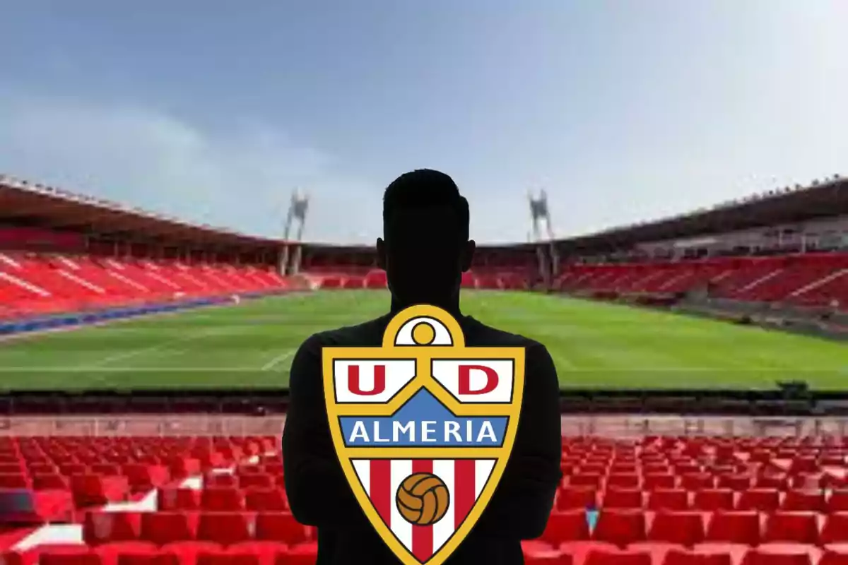 Persona en silueta sosteniendo el escudo del equipo de fútbol UD Almería en un estadio vacío con asientos rojos.