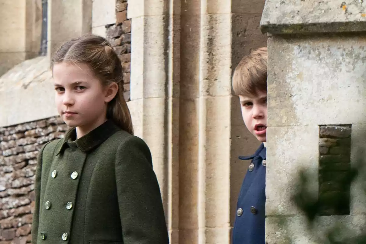 La princesa Charlotte y el príncipe Louis con abrigos elegantes están de pie junto a una pared de piedra.