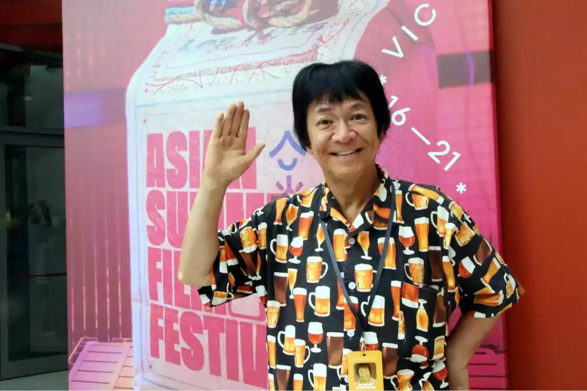 Un home somrient amb una camisa estampada de cerveses aixeca la mà davant d'un cartell de l'Asian Summer Film Festival.