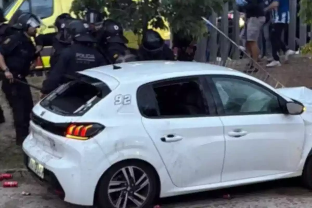 Un cotxe blanc amb el vidre del darrere trencat està estacionat a prop d'un grup de policies amb equip antidisturbis. Un cotxe blanc amb el vidre del darrere trencat està estacionat a prop d'un grup de policies amb equip antidisturbis.
