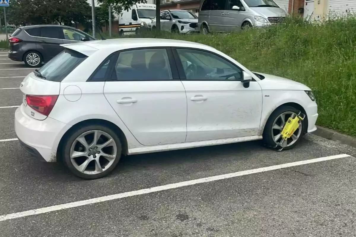 Un coche blanco estacionado en un aparcamiento con un cepo amarillo en la rueda delantera derecha.