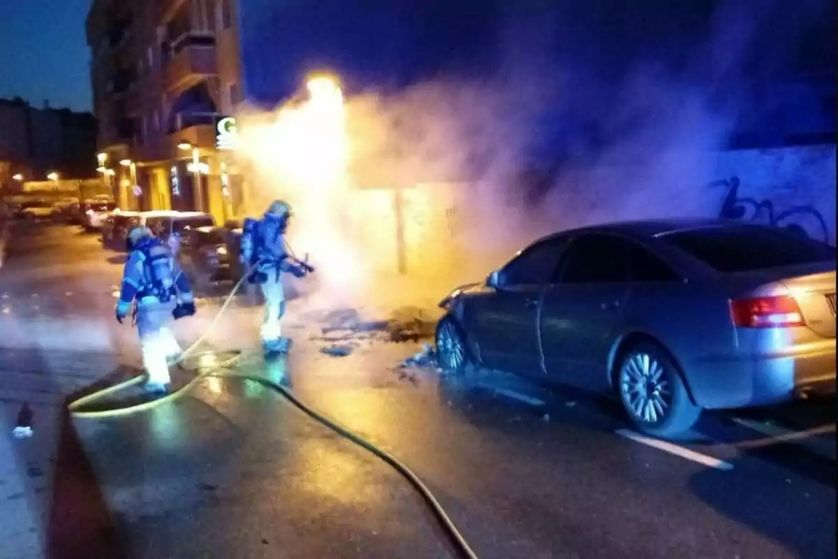 Bombers apagant un incendi en un cotxe estacionat en un carrer durant la nit.