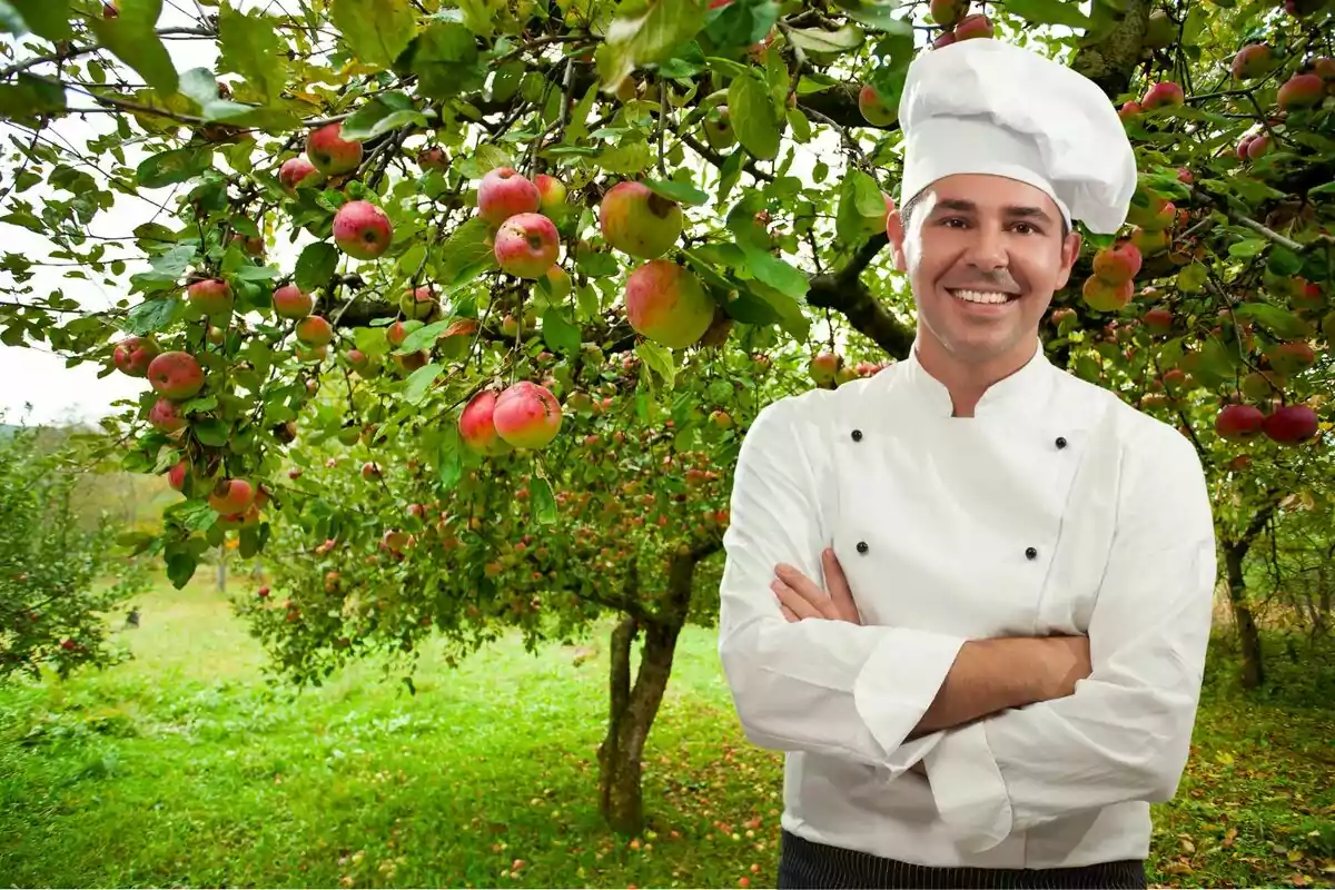 Xef somrient amb uniforme blanc de peu davant d'un arbre de pomes en un hort.