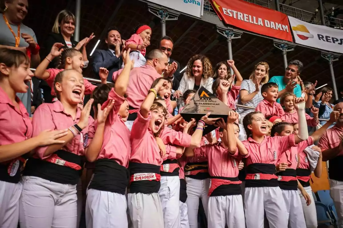 Un grup de persones celebra amb entusiasme mentre sostenen un trofeu en un esdeveniment, tothom vesteix camises rosades i pantalons blancs, envoltats d'espectadors que aplaudeixen.