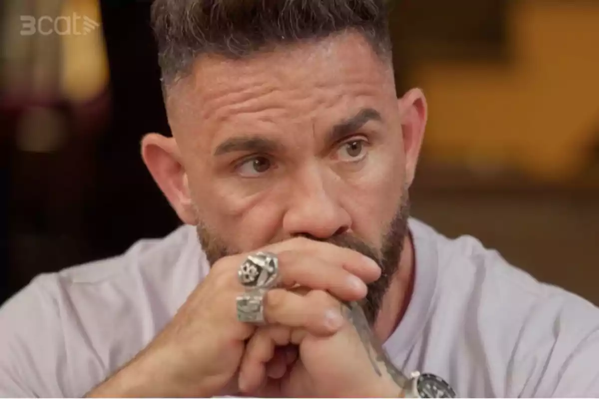 Un hombre con barba y cabello corto, con las manos juntas frente a su boca, lleva varios anillos y parece pensativo.