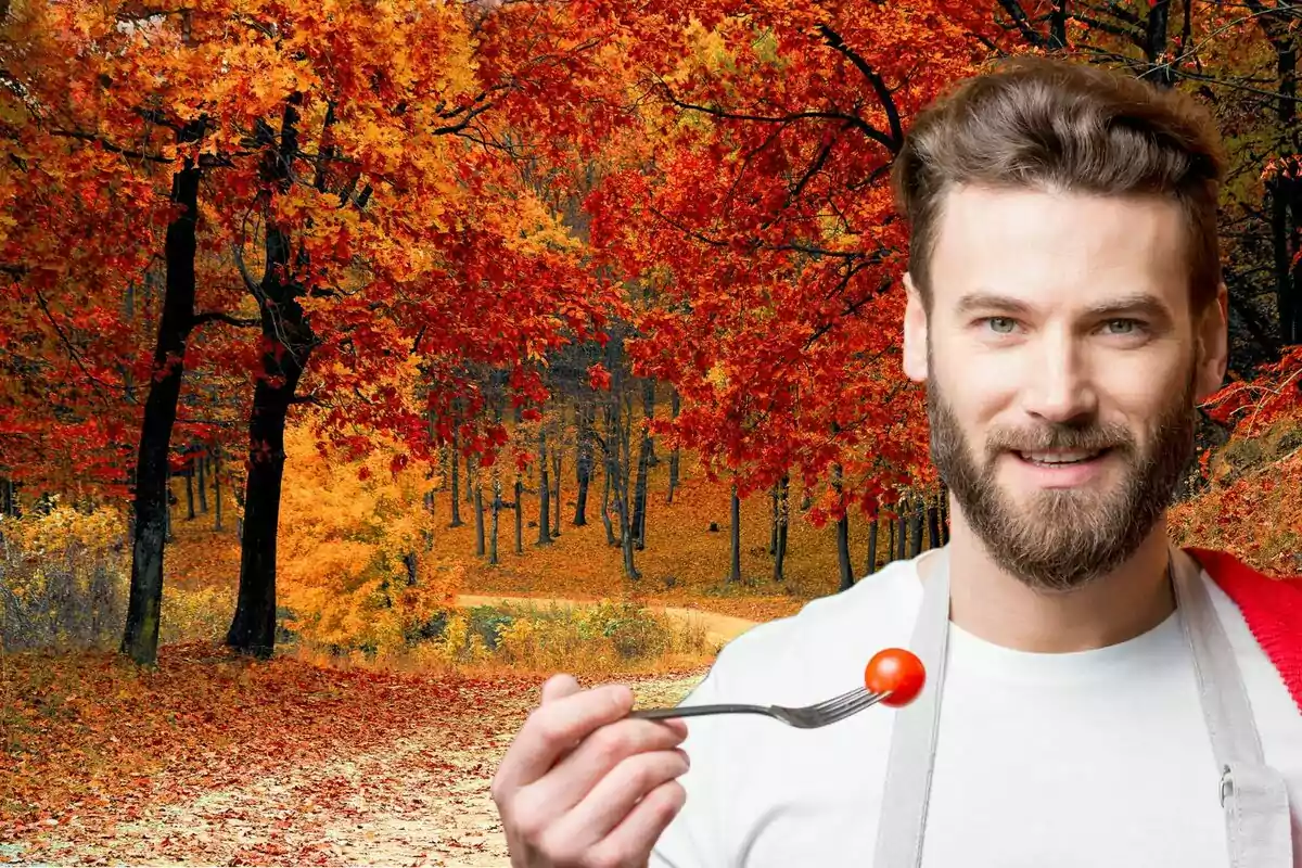 Un home amb barba sosté una forquilla amb un tomàquet cherry davant d'un paisatge tardorenc amb arbres de fulles vermelles i taronges.