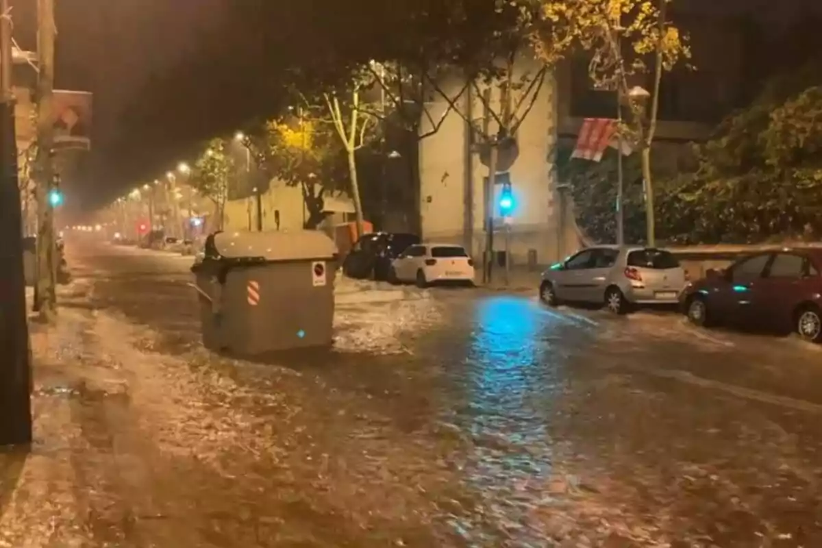 Calle inundada por la lluvia con contenedor flotando y coches estacionados a los lados.