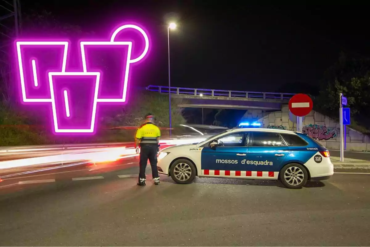 Un agent de policia de trànsit al costat d'un cotxe patrulla dels Mossos d'Esquadra en una carretera a la nit, amb un rètol de neó rosa en forma de gots al fons.