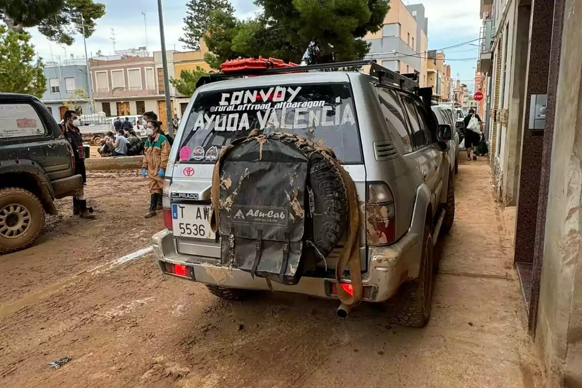 Un vehicle tot terreny amb rètols a la finestra del darrere que diu "Convoy Ajuda València" està estacionat en un carrer de terra, amb persones al voltant i edificis al fons.