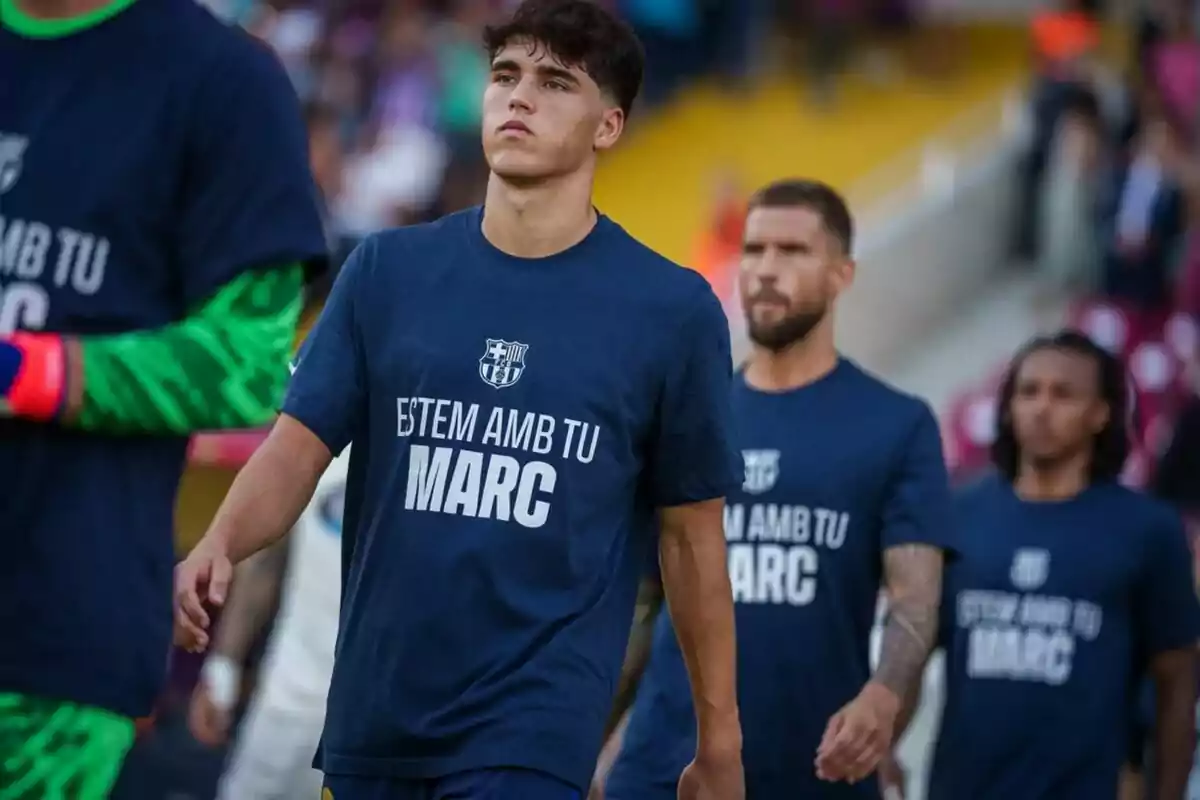 Jugadors de futbol caminant al camp amb samarretes blaves que diuen “ESTEM AMB EL TEU MARC”.