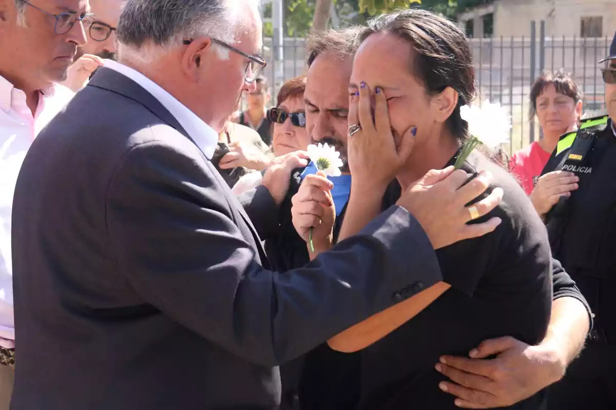 Persones consolant-se mútuament mentre sostenen flors blanques en un entorn exterior.