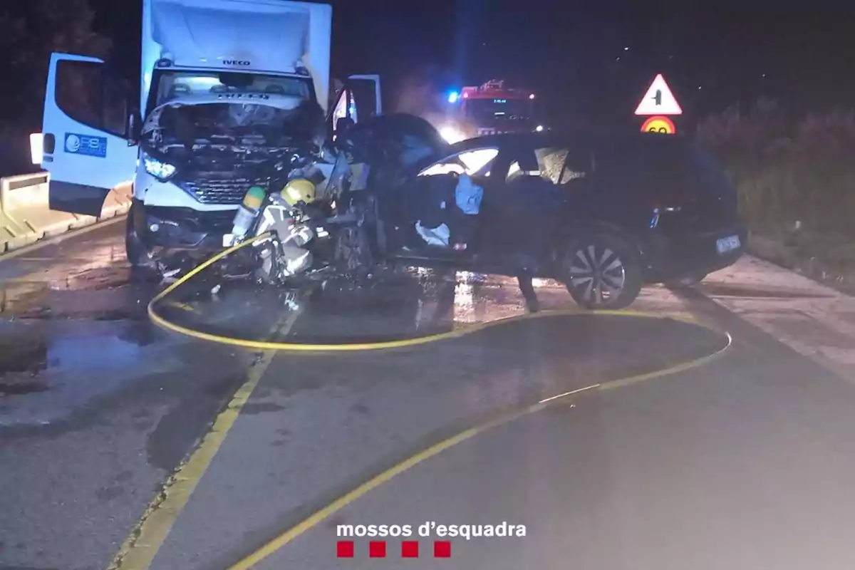 Bombers treballant a l'escena d'un accident entre un camió i un cotxe en una carretera, amb vehicles d'emergència i senyals de trànsit visibles al fons. Bombers treballant a l'escena d'un accident entre un camió i un cotxe en una carretera, amb vehicles d'emergència i senyals de trànsit visibles al fons.