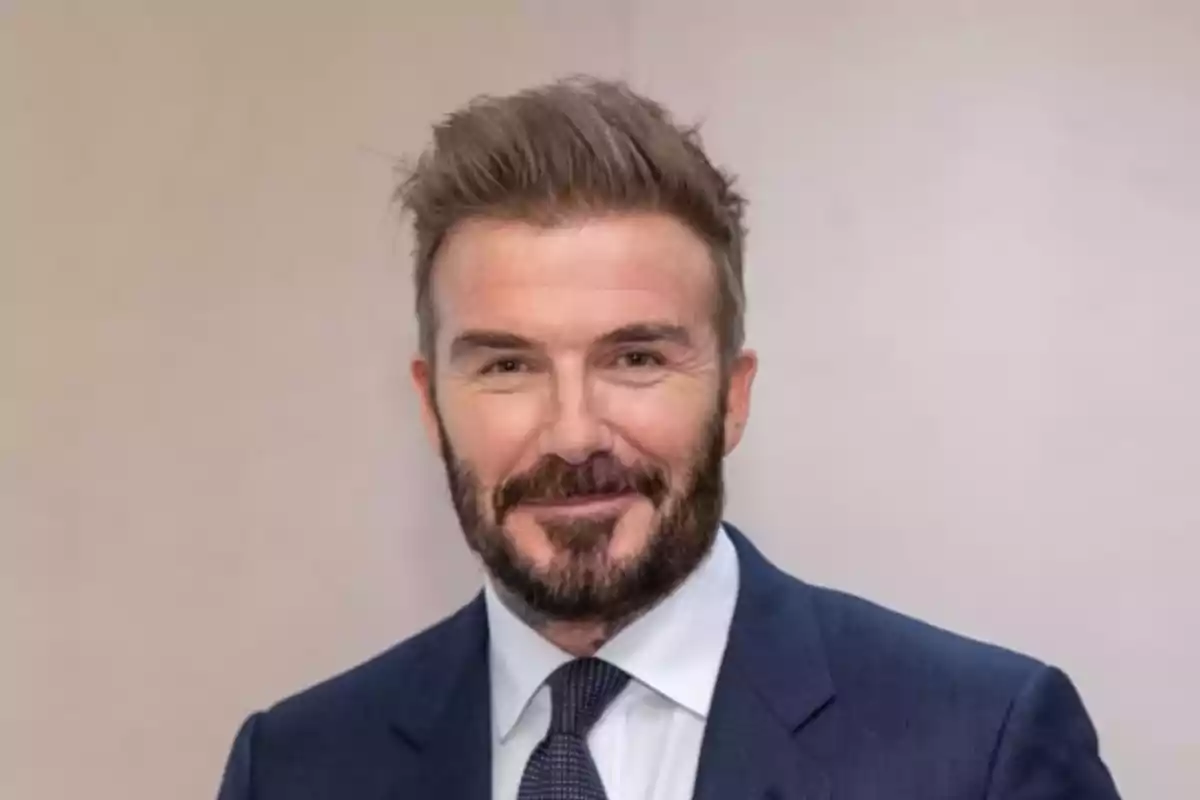 A man with a beard and short hair dressed in a suit and tie smiles in front of a neutral background. A man with a beard and short hair dressed in a suit and tie smiles in front of a neutral background.