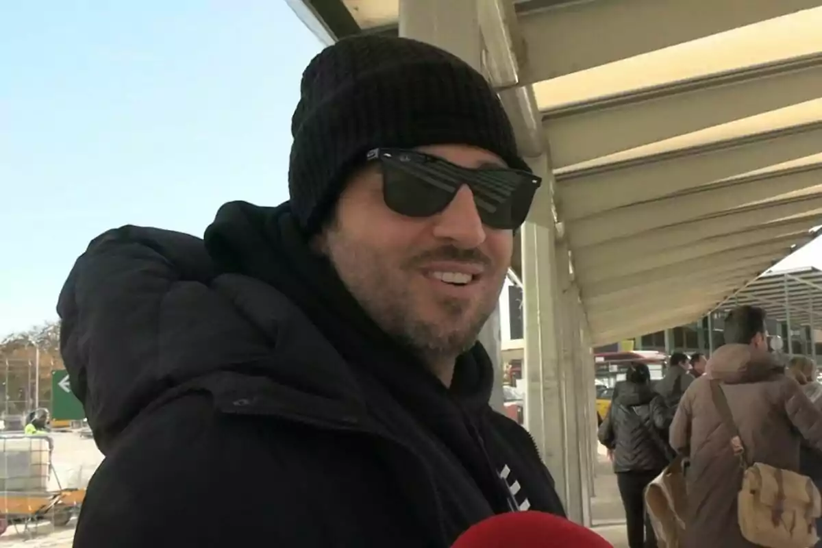 David Bustamante con gafas de sol y gorro negro sonríe mientras está al aire libre en un lugar concurrido. David Bustamante con gafas de sol y gorro negro sonríe mientras está al aire libre en un lugar concurrido.