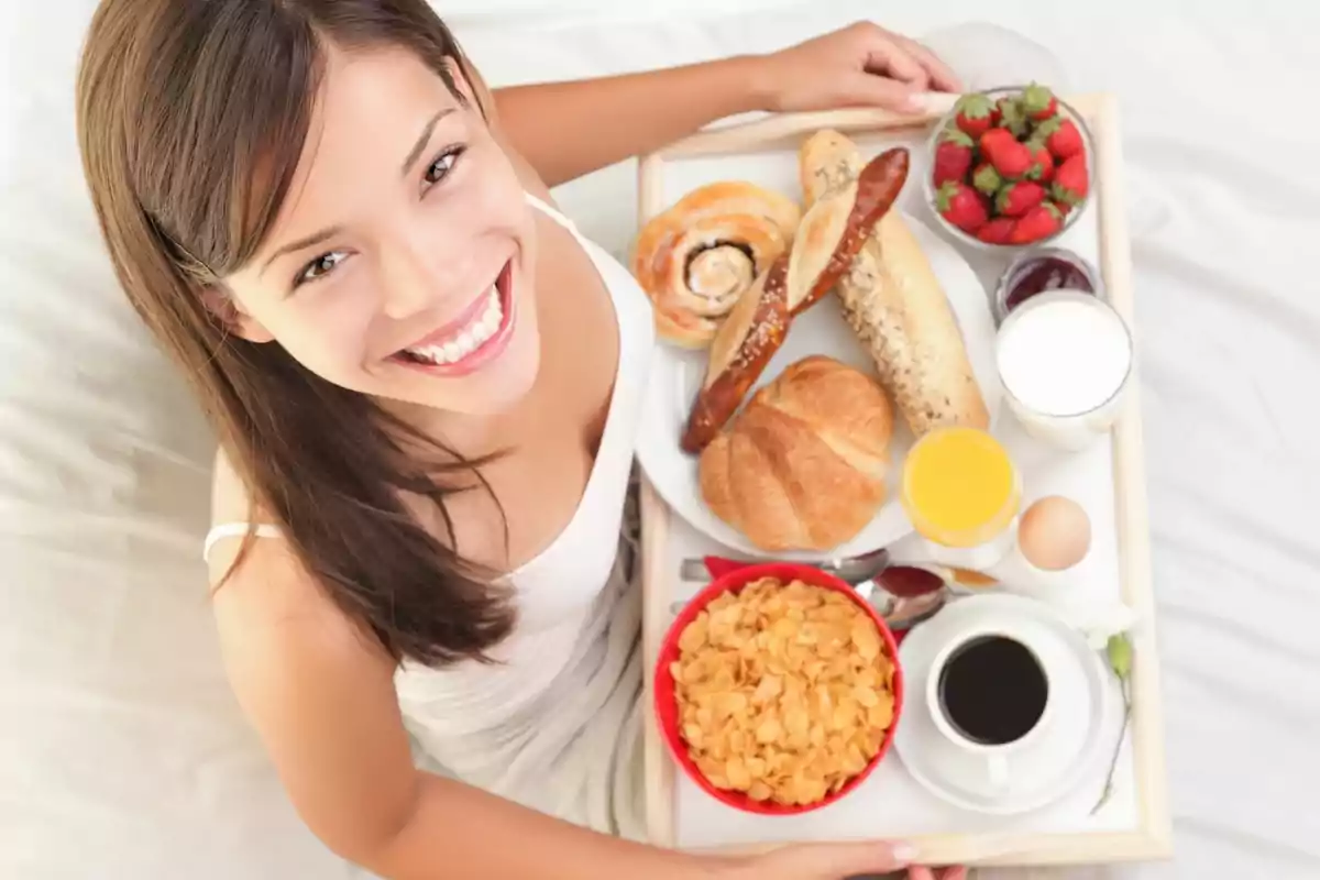 Mujer sonriente disfrutando de un desayuno en la cama con una bandeja que incluye croissants, pan, cereales, café, jugo de naranja, leche, fresas y un huevo.