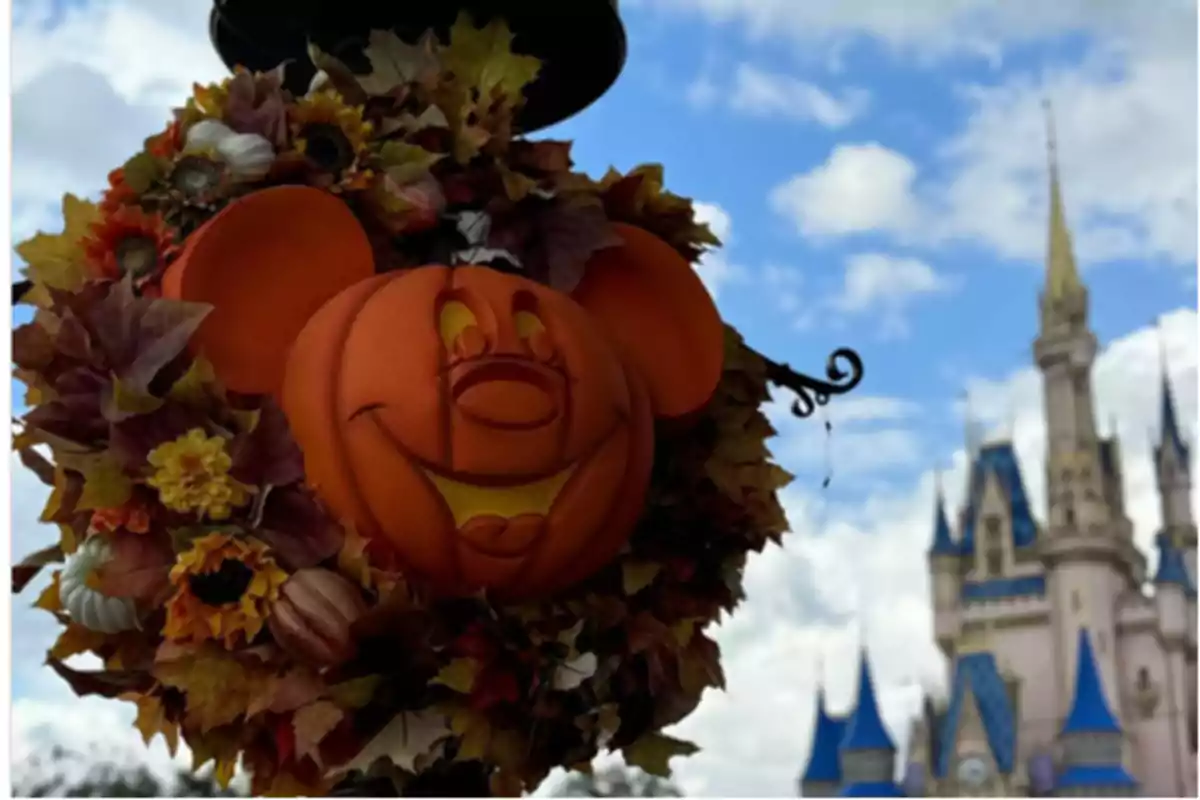 Decoración de Halloween con una calabaza sonriente y orejas de ratón rodeada de hojas otoñales, con un castillo de fondo bajo un cielo azul.