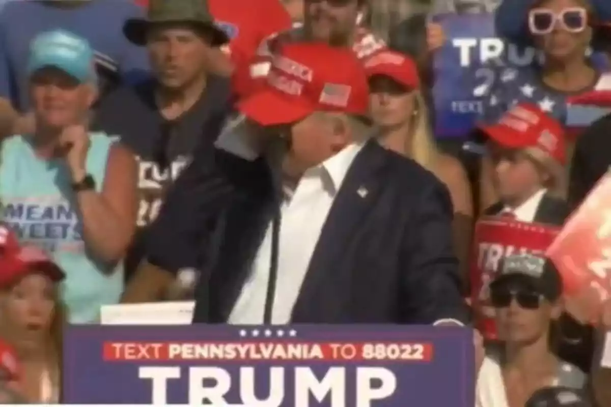 Un home amb una gorra vermella està parlant en un míting polític, envoltat de persones que porten gorres i samarretes amb missatges de suport.