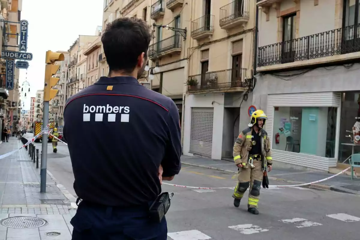 Un bomber d?esquena observa un altre bomber caminant per un carrer acordonat en una ciutat.