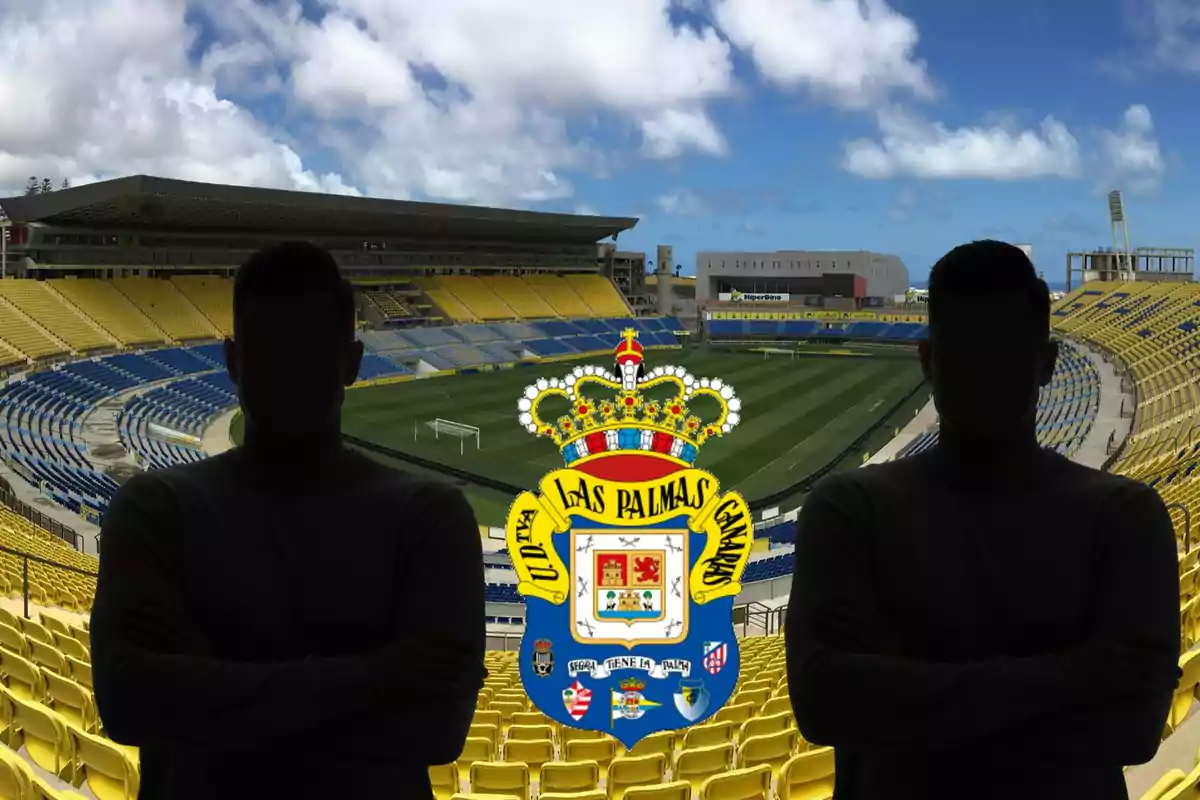 Dues persones amb siluetes fosques posen davant d'un estadi de futbol buit amb seients grocs i blaus, amb l'escut de l'equip de futbol UD Las Palmas al centre de la imatge.