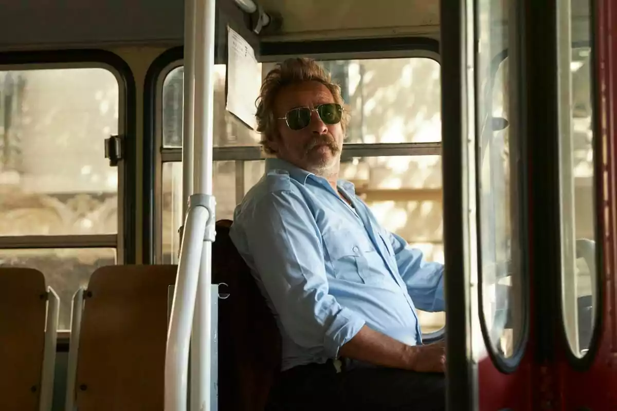 Un hombre con gafas de sol y camisa azul está sentado al volante de un autobús.