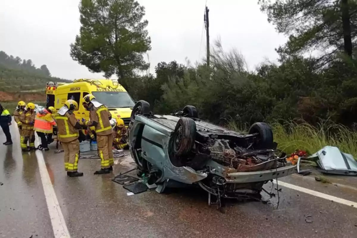 Accidente de tráfico con un coche volcado en una carretera mojada mientras bomberos y personal de emergencia trabajan en el lugar.