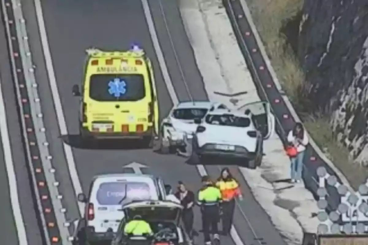 Una ambulancia y varios vehículos están detenidos en una carretera mientras personas, incluyendo personal de emergencia, se encuentran alrededor.