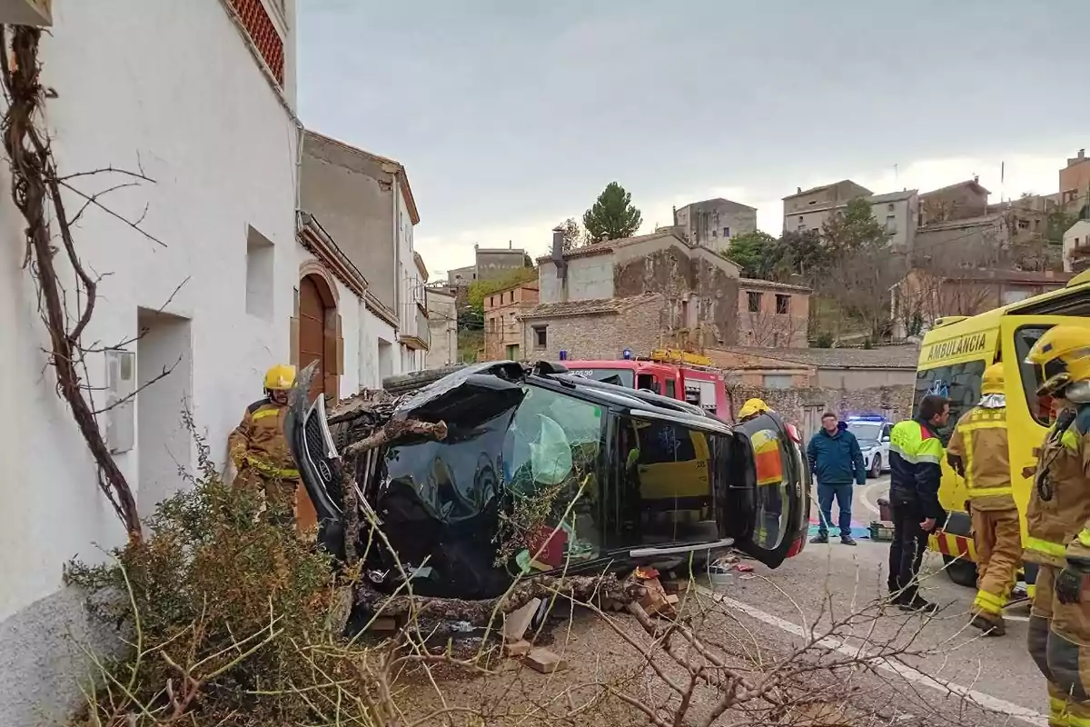 Un coche volcado en una calle con bomberos y personal de emergencia alrededor, junto a una ambulancia y un camión de bomberos.