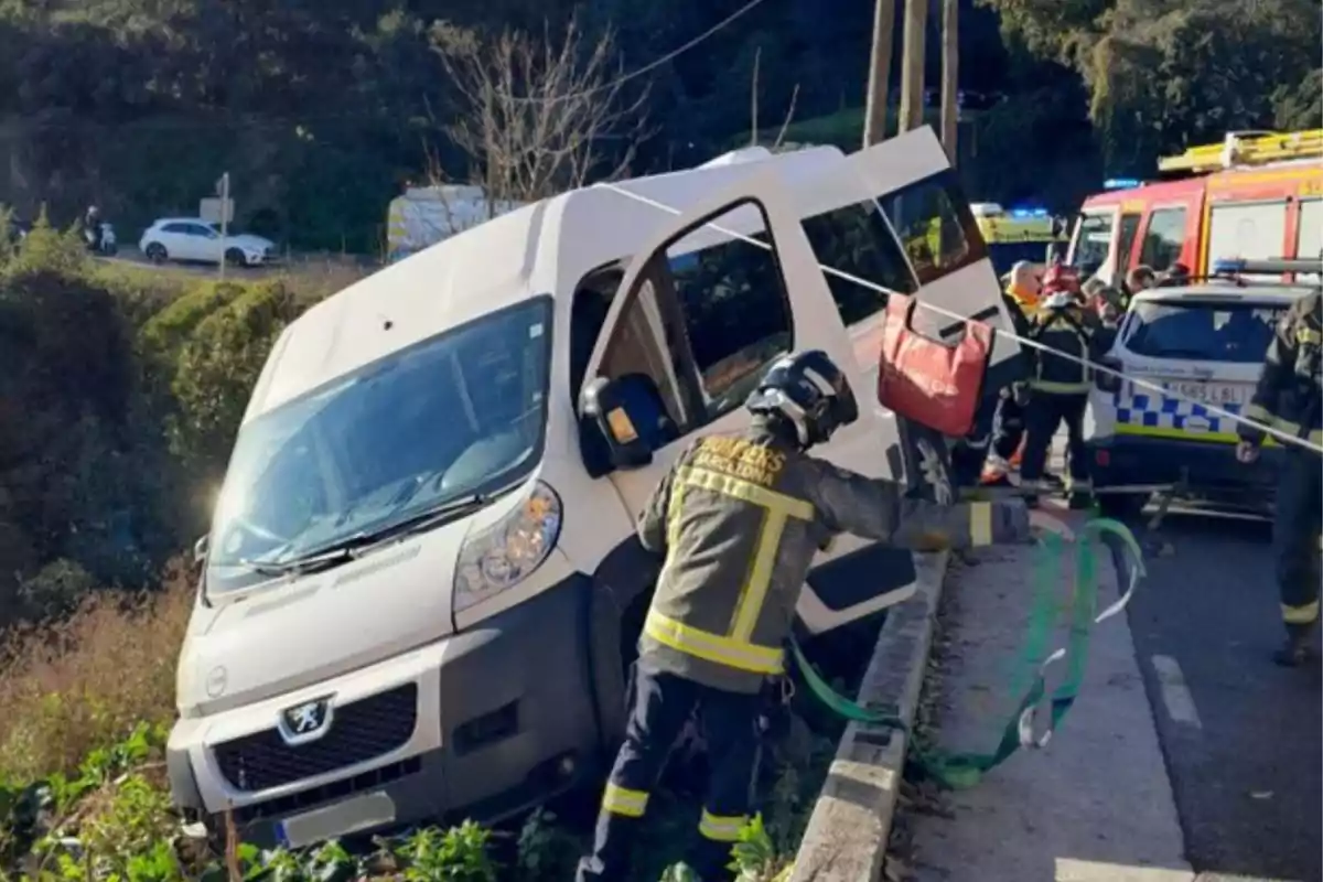 Un grupo de bomberos trabaja para asegurar una furgoneta que ha quedado inclinada en el borde de una carretera mientras varios vehículos de emergencia están presentes en el lugar.