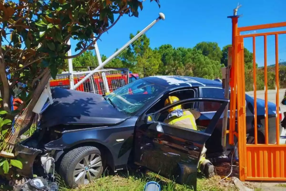 Un coche negro se ha estrellado contra una valla y un árbol, con un bombero trabajando en la escena.