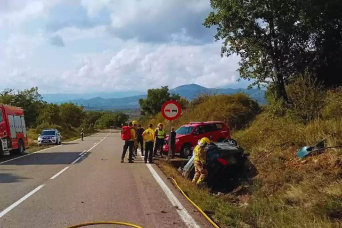 Un grup de bombers i rescatistes atenen un accident de trànsit en una carretera rural, amb un cotxe bolcat a la cuneta i vehicles d?emergència al?escena.