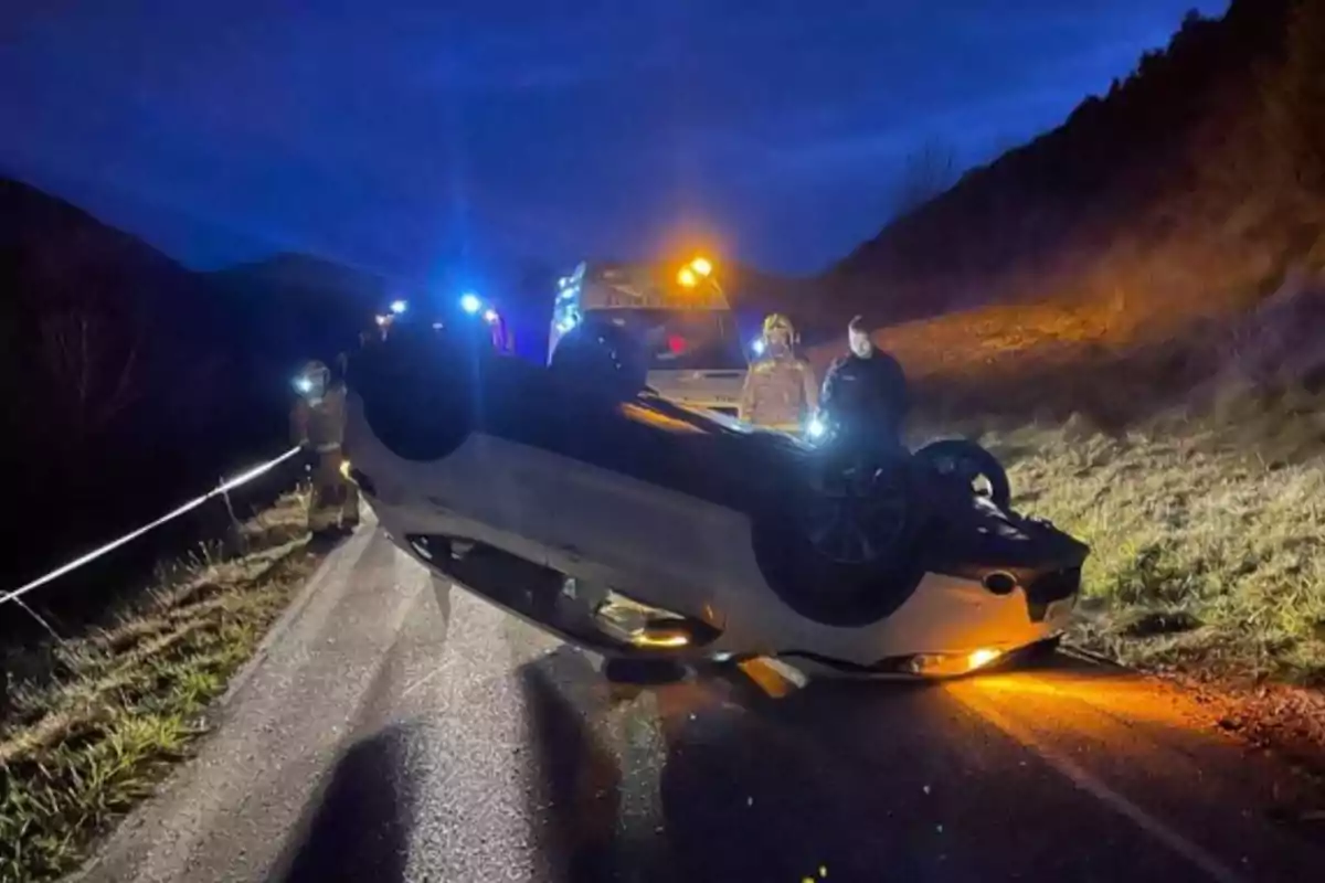 Un coche volcado en una carretera con luces de emergencia y personas alrededor en la noche.