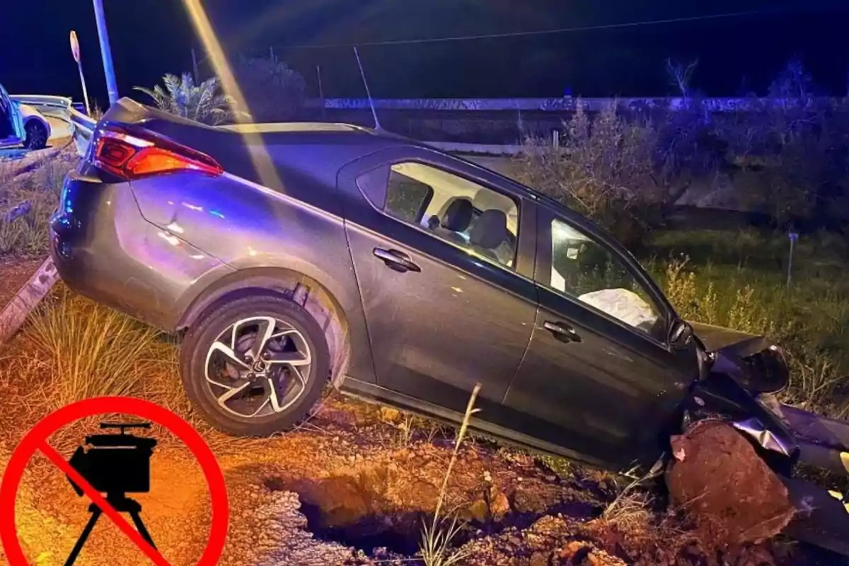 Un cotxe gris accidentat en una rasa al costat de la carretera durant la nit amb llums d'emergència il·luminant l'escena i un símbol de prohibició sobre un radar de velocitat a la cantonada inferior esquerra.