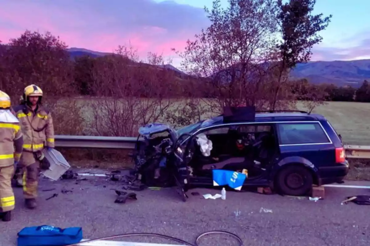 Un cotxe blau amb la part davantera danyada després d'un accident en una carretera rural al capvespre amb dos bombers a l'escena.