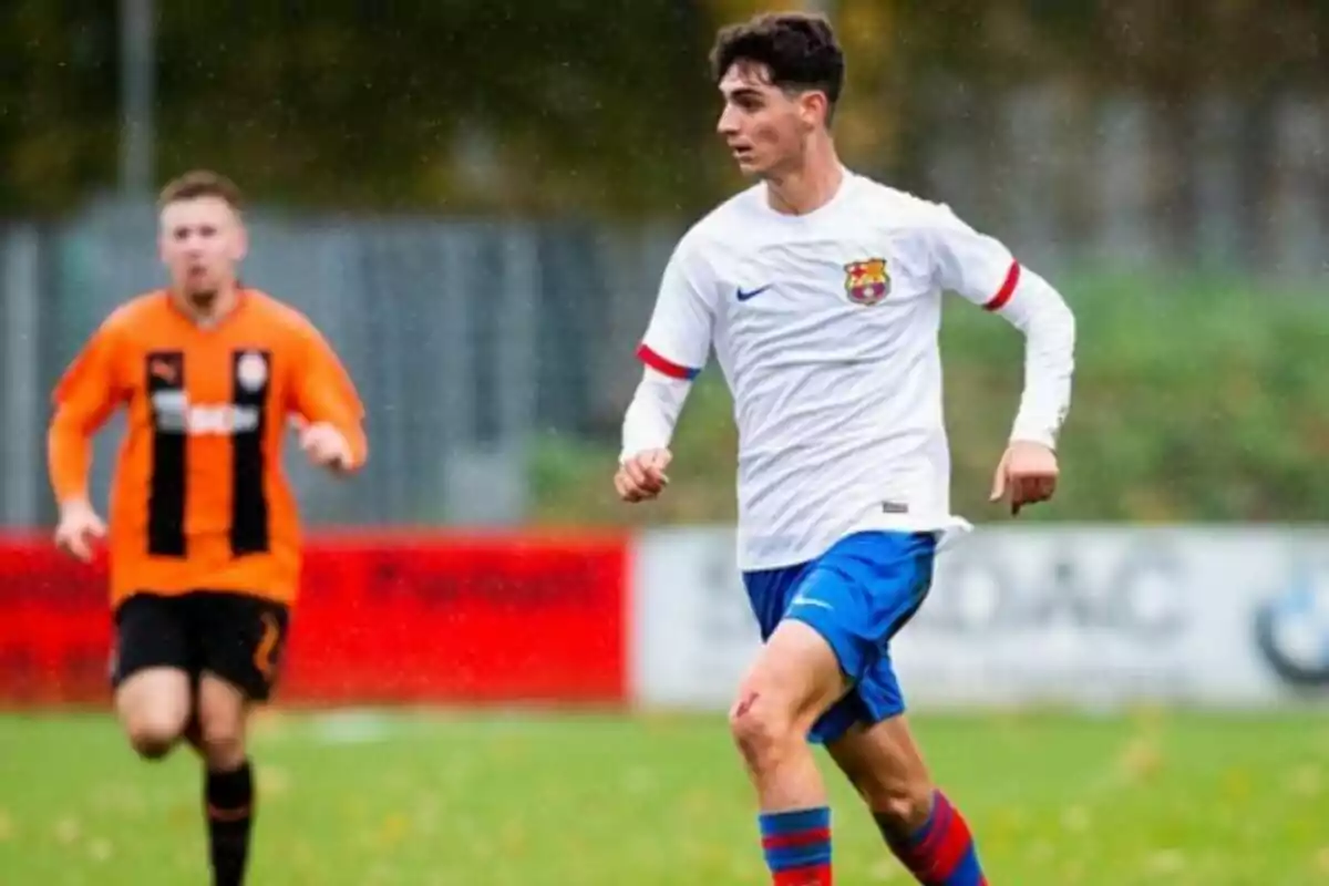 Dos jugadors de futbol en un camp, un amb uniforme blanc i blau del FC Barcelona i un altre amb uniforme taronja i negre.