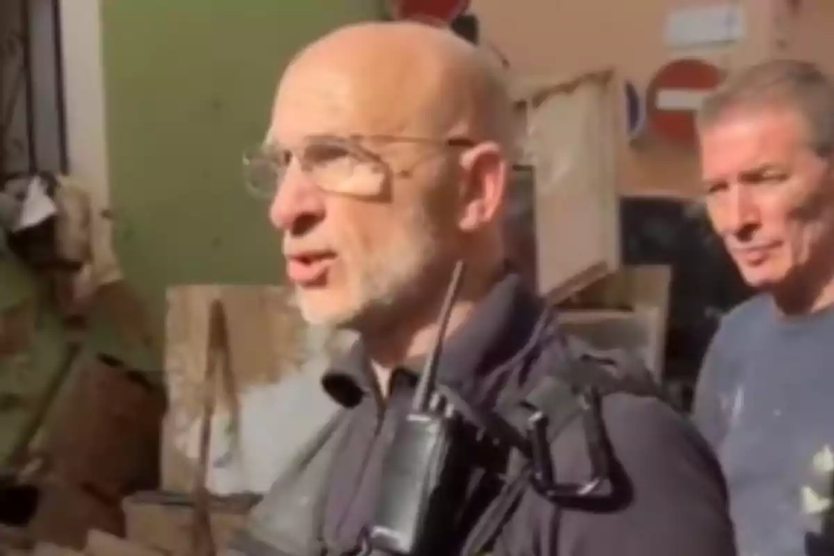 Un hombre con gafas y radio en el hombro está hablando al aire libre, acompañado de otra persona.