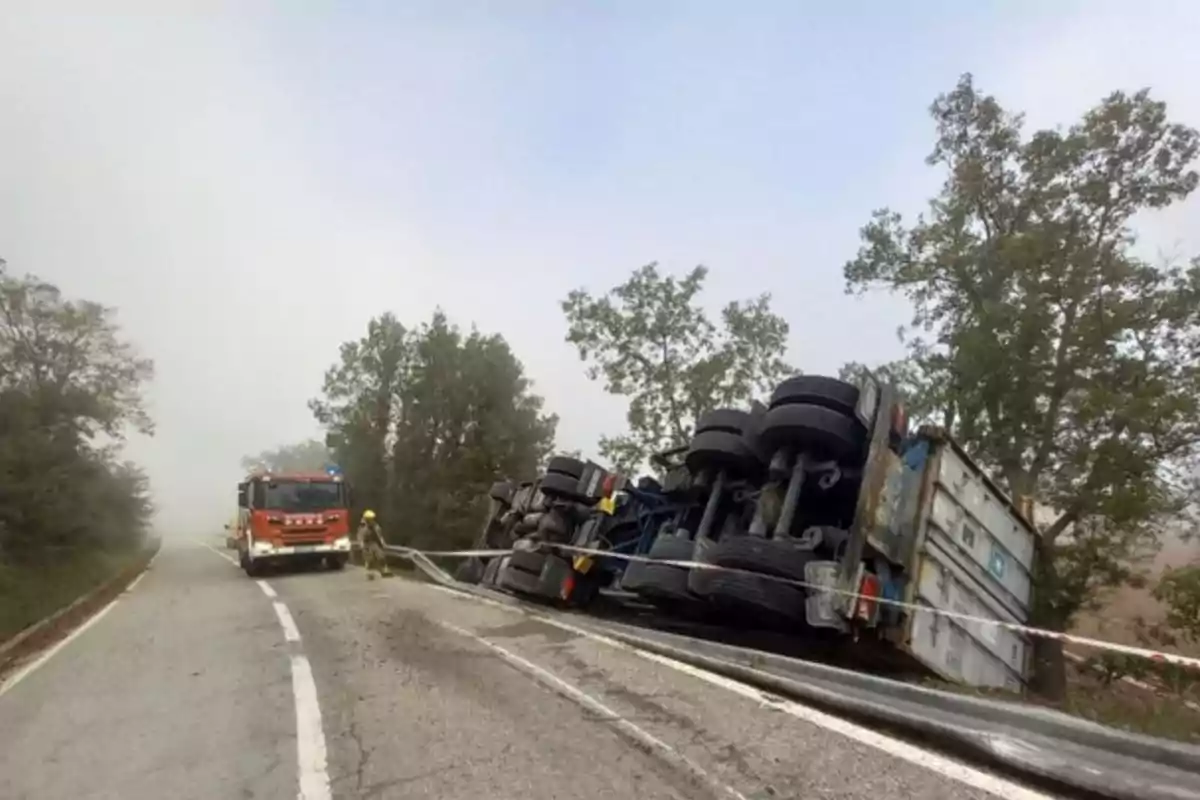 Un camió bolcat a la vora d'una carretera amb un camió de bombers i un bomber a l'escena.