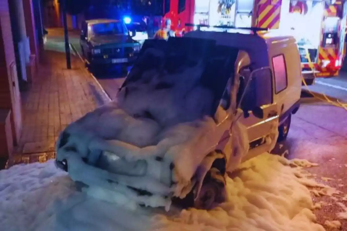 Un vehículo cubierto de espuma en una calle, con luces de emergencia y un camión de bomberos al fondo.