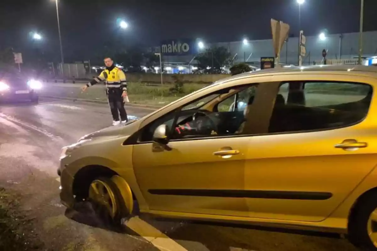 Un coche con una rueda dañada en una carretera por la noche mientras un oficial de policía señala hacia el vehículo.