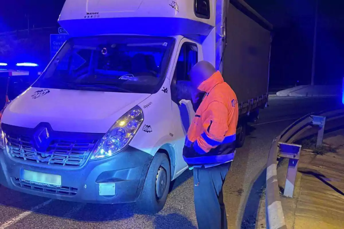 Un hombre con chaqueta reflectante naranja está de pie junto a una furgoneta blanca en una carretera iluminada por luces azules durante la noche.