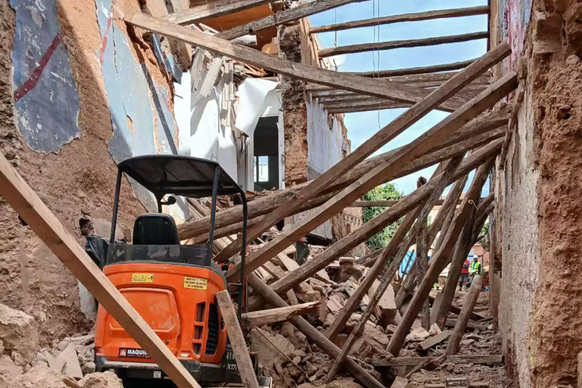 Una excavadora taronja es troba en un edifici en ruïnes amb runes i bigues de fusta caigudes.