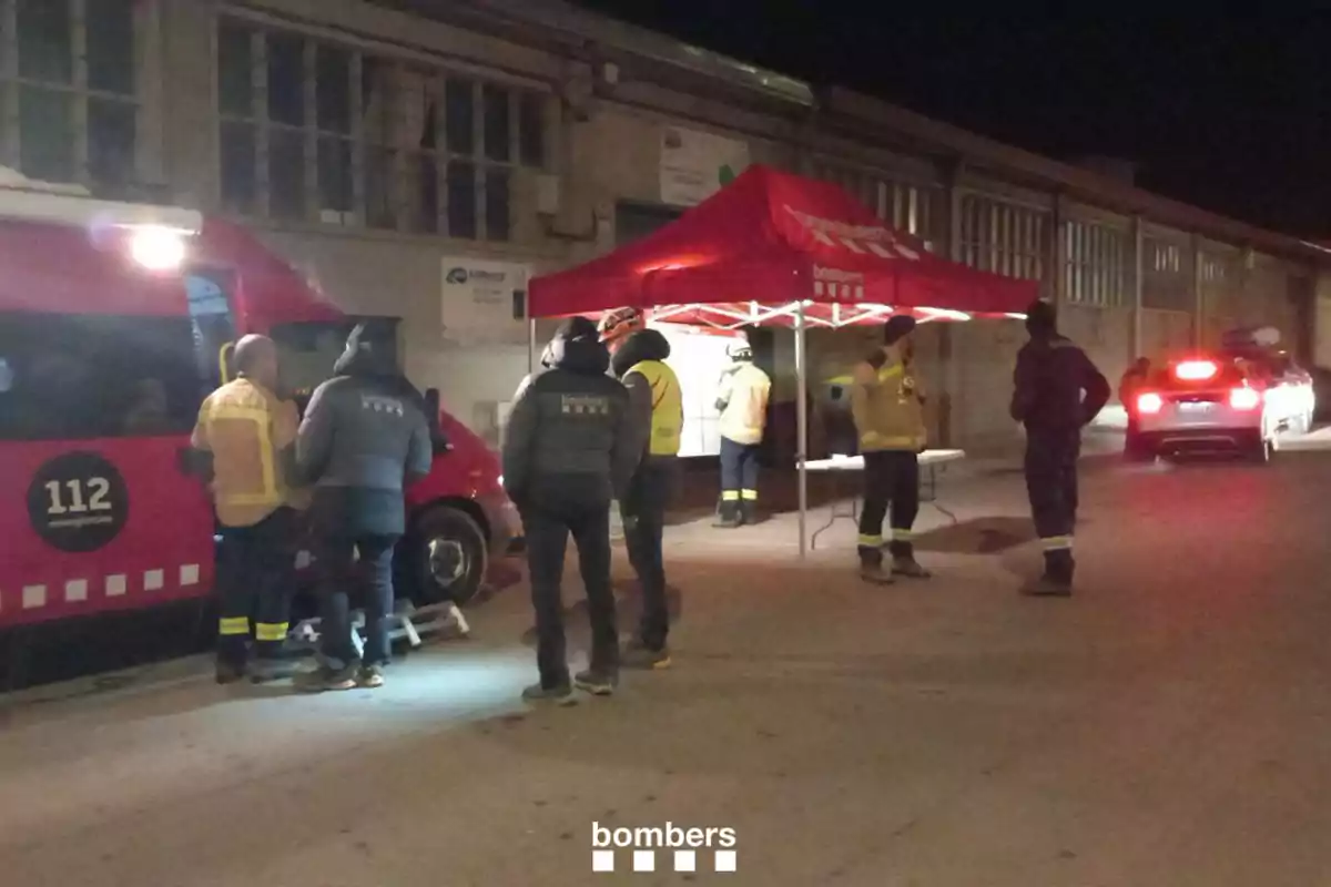 Un grup de bombers es troben reunits al costat d'una furgoneta vermella i una carpa en una zona industrial durant la nit.