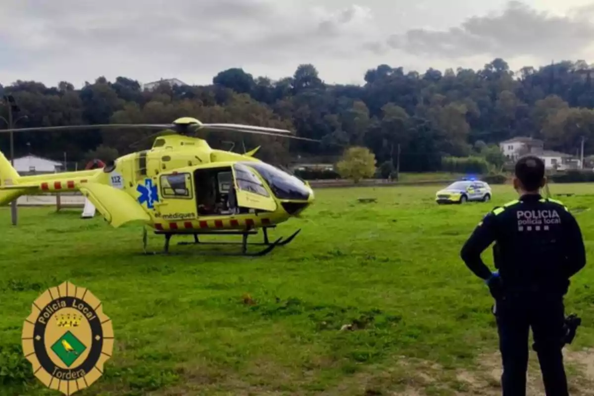 Un helicòpter groc d?emergències mèdiques està estacionat en un camp d?herba amb un oficial de policia local observant i un cotxe patrulla al fons.