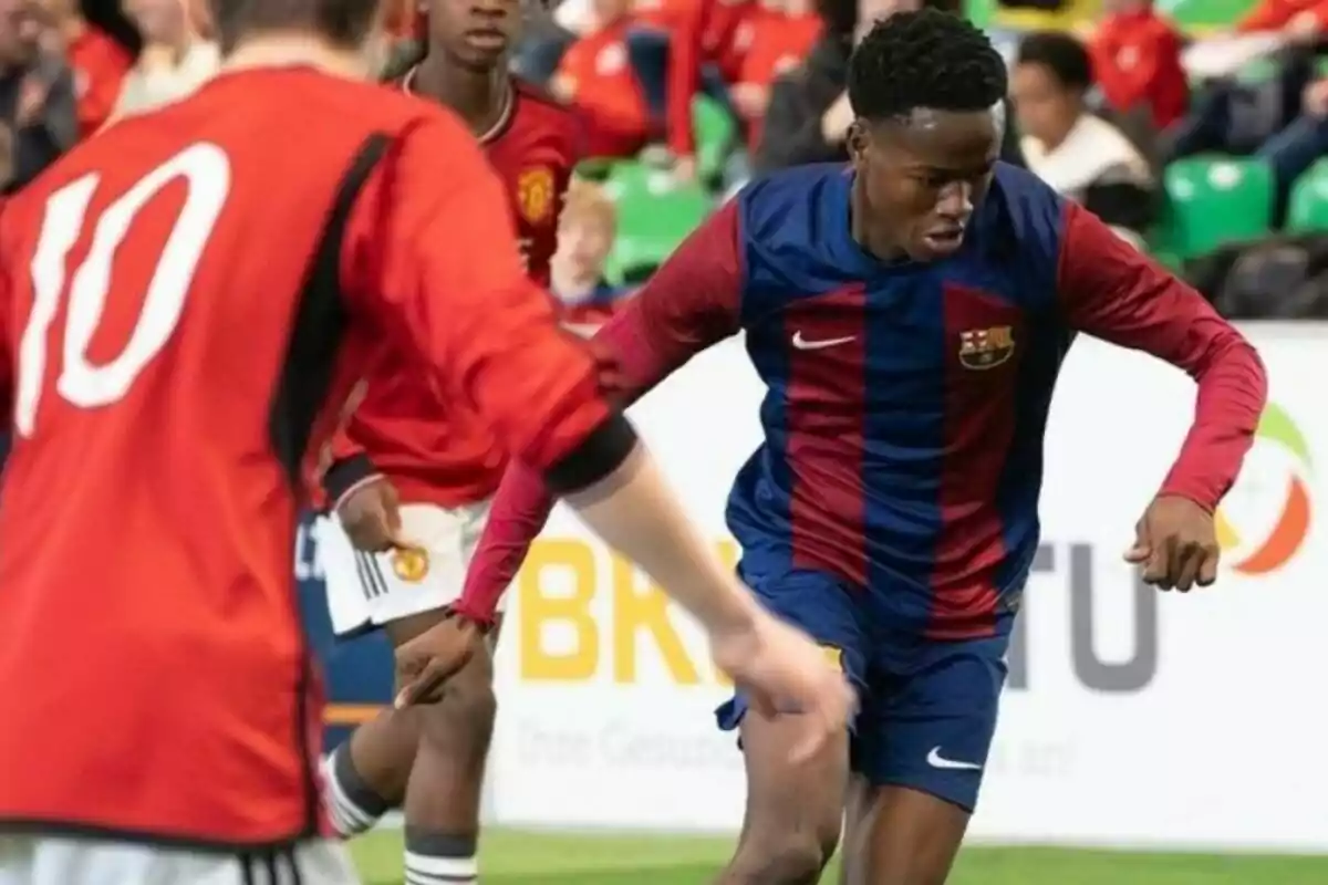 Jugadors de futbol en acció durant un partit, un amb uniforme vermell i un altre amb uniforme blau i granat.