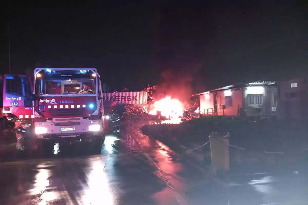 Un camión de bomberos está estacionado en una carretera mojada durante la noche mientras un incendio arde intensamente cerca de un edificio y un contenedor.